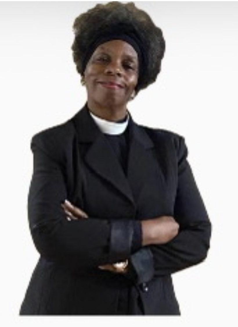 Portrait of Pastor Barbara Lewis-Pierce, a Black woman with voluminous curly dark hair held back by a black headband, wearing a black blazer over a black shirt with a white clerical collar, arms crossed, gold watch on left wrist, plain off-white studio background